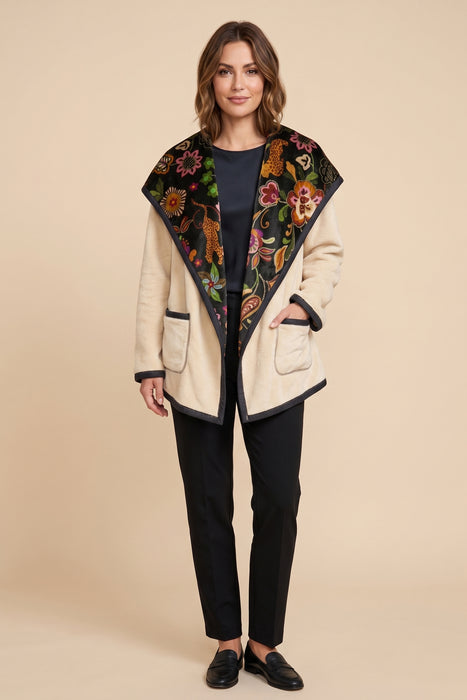 A woman modeling a cream-colored wrap jacket with a large black floral embroidered collar, paired with black pants and black loafers, against a solid beige background.