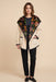 A woman modeling a cream-colored wrap jacket with a large black floral embroidered collar, paired with black pants and black loafers, against a solid beige background.
