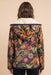 A woman viewed from the back wearing a hooded jacket with a dark base covered in a dense multicolor floral and animal print, with a cream sherpa collar, against a solid beige background.
