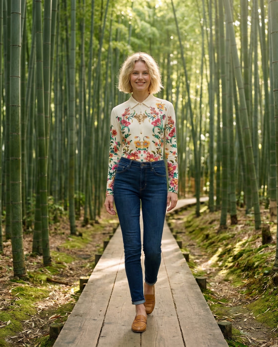 A woman walking on a wooden boardwalk path through a dense bamboo forest, wearing a cream long-sleeve button-up shirt with a colorful floral and butterfly print, dark blue jeans, and tan boots.
