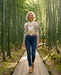 A woman walking on a wooden boardwalk path through a dense bamboo forest, wearing a cream long-sleeve button-up shirt with a colorful floral and butterfly print, dark blue jeans, and tan boots.