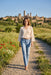 A woman walking on a gravel path in a sunny rural landscape, wearing a cream lace long-sleeve blouse and light blue jeans, with a hillside town and towers in the background.