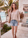 A woman stands on a tropical beachside patio wearing a light beige knit long-sleeve top, distressed denim shorts, and brown sandals, carrying a woven shoulder bag.