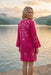 A woman stands by a lake at sunset wearing a bright pink embroidered tunic dress with white floral and botanical patterns. The background shows calm water, distant hills, and a soft golden sky.