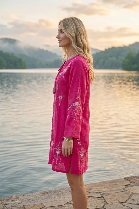 A woman stands in profile by a calm lake at sunset, wearing a bright pink embroidered tunic or cover-up dress with white floral detailing.