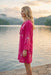 A woman stands in profile by a calm lake at sunset, wearing a bright pink embroidered tunic or cover-up dress with white floral detailing.