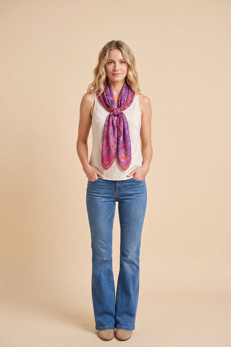 A woman modeling a vibrant purple and pink patterned silk scarf tied around her neck, paired with a white sleeveless top and blue jeans, against a solid beige studio background.