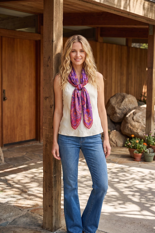 A woman stands on a rustic wooden porch wearing a cream sleeveless top, blue jeans, and a vibrant purple and pink patterned silk scarf tied around her neck.
