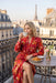 A woman sits on a Parisian balcony wearing a vibrant red floral robe, holding a teacup. A small table next to her holds croissants and a vase of flowers, with the Eiffel Tower and Parisian rooftops in the background.