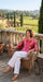A woman sits in a wicker chair on a stone terrace, wearing a pink and red patterned long-sleeve top with white wide-leg pants. She holds a glass of white wine, with a vineyard and rustic buildings in the background.