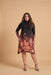A woman modeling a long-sleeve mock-neck midi dress with a black base and a large floral print in warm coral, orange, and pink tones that fade into a darker pattern near the hem, set against a solid beige studio background.
