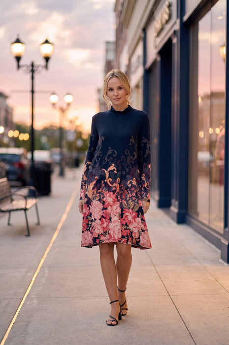 A woman stands on a city sidewalk at dusk wearing a long-sleeve mock-neck midi dress with a dark navy base and a pink, red, and orange floral print concentrated at the hem and sleeves. The background shows blurred storefronts, street lamps, and parked cars.