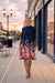 A woman stands on a city sidewalk at dusk wearing a long-sleeve mock-neck midi dress with a dark navy base and a pink, red, and orange floral print concentrated at the hem and sleeves. The background shows blurred storefronts, street lamps, and parked cars.