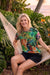 A woman sitting in a hammock outdoors wearing a colorful tropical print t-shirt with tiger and palm tree designs, paired with black shorts, surrounded by lush green tropical foliage.