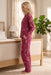 A woman stands in profile in a bedroom wearing a long-sleeve floral pajama set in a deep pink/magenta color. The set includes a button-front top and matching pants with a small multicolor floral print.