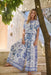 A woman models a white short-sleeve maxi dress with a blue floral toile pattern, standing on stone steps outdoors next to a tree trunk.