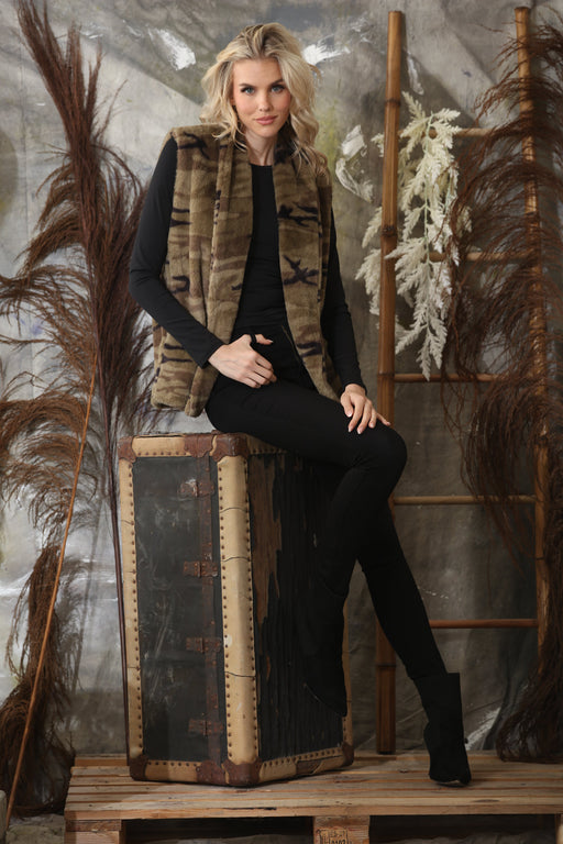 Woman wearing a Johnny Was Workshop Molly Camo Faux Fur Vest in a camouflage pattern, styled with black outfit and seated in a rustic indoor setting.