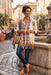 A woman stands in a European-style street setting wearing a cream-colored bohemian tunic shirt with colorful blue, orange, and green floral patterns, paired with dark blue jeans and tan ankle boots.