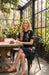 A woman sits at a rustic wooden table inside a sunlit greenhouse, wearing a black short-sleeve dress with colorful floral embroidery and brown sandals.