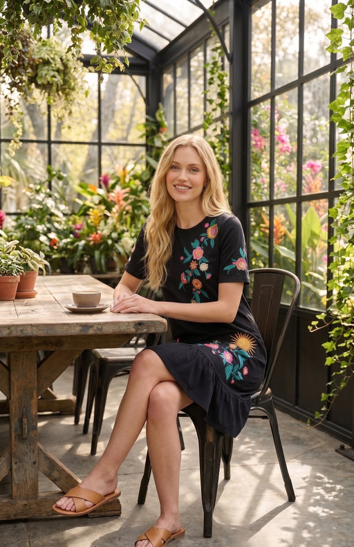 A woman sits at a rustic wooden table inside a sunlit greenhouse, wearing a black short-sleeve dress with colorful floral embroidery and brown sandals.