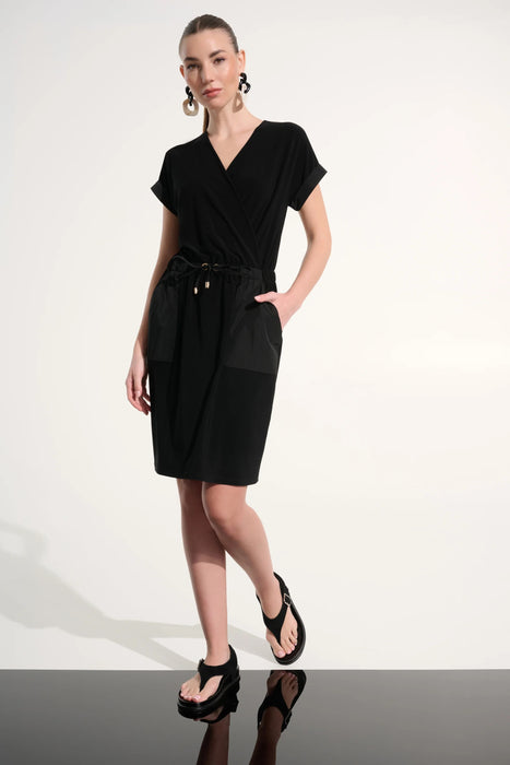 Woman wearing Joseph Ribkoff black silky knit and memory straight wrap dress, featuring a wrap neckline, drawstring waist, short sleeves, and a sleek, comfortable fit with pockets.