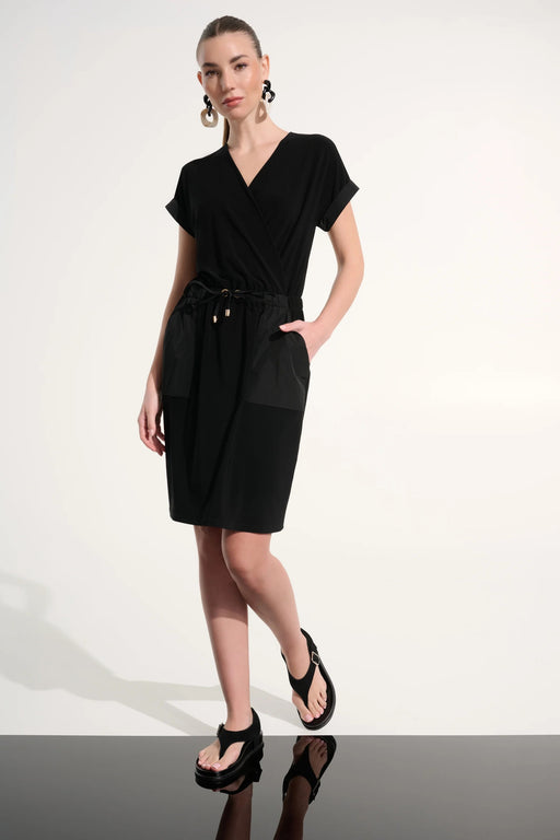 Woman wearing Joseph Ribkoff black silky knit and memory straight wrap dress, featuring a wrap neckline, drawstring waist, short sleeves, and a sleek, comfortable fit with pockets.