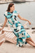 Woman wearing Joseph Ribkoff green multi geometric print fit and flare dress with cap sleeves and cinched waist, offering a modern and elegant silhouette.