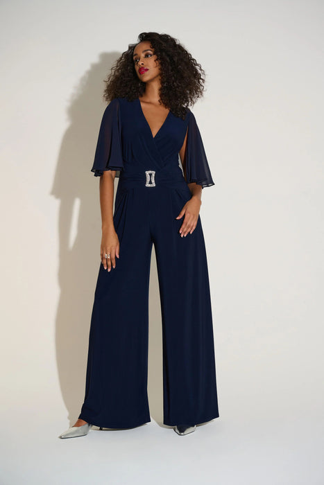 Woman wearing a navy blue wide leg jumpsuit with chiffon flutter cape sleeves and rhinestone buckle detail
