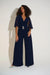 Woman wearing a navy blue wide leg jumpsuit with chiffon flutter cape sleeves and rhinestone buckle detail