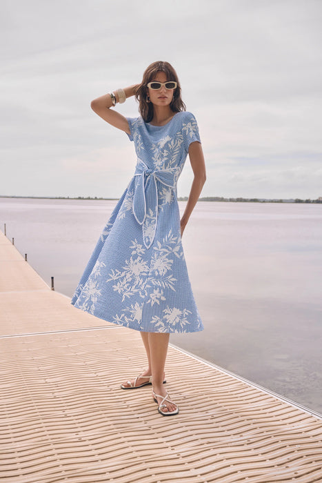Woman wearing a Joseph Ribkoff vanilla and blue floral print fit and flare midi dress with short sleeves and a tie waist, styled for a chic summer look