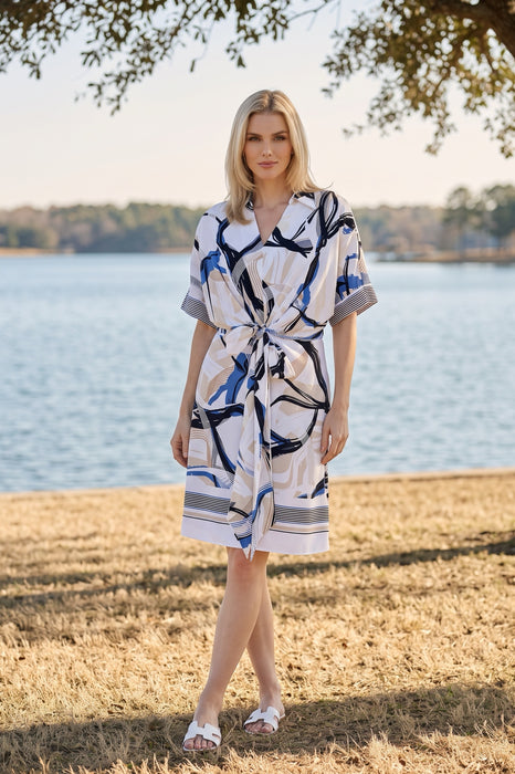 A woman wearing a short-sleeve white wrap-style dress with an abstract blue and gray stripe prints from Joseph Ribkoff