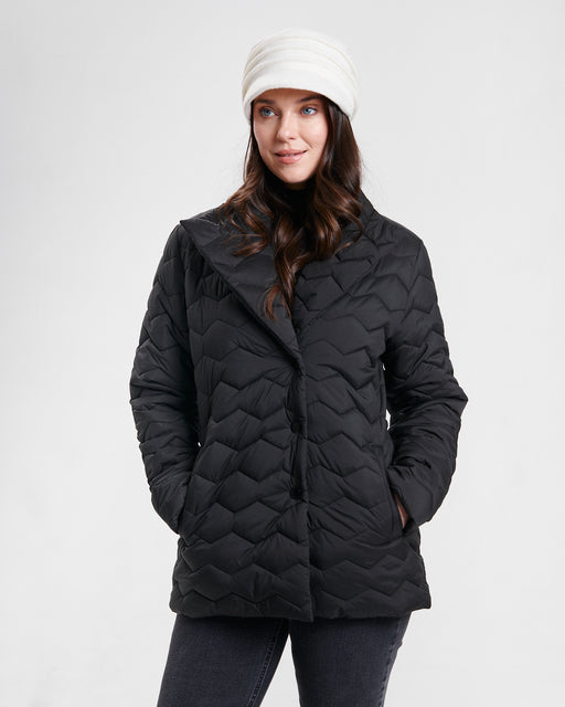 A woman wearing a black quilted puffer jacket with a wide collar, paired with a white knit beanie and dark jeans, photographed against a light gray studio background.