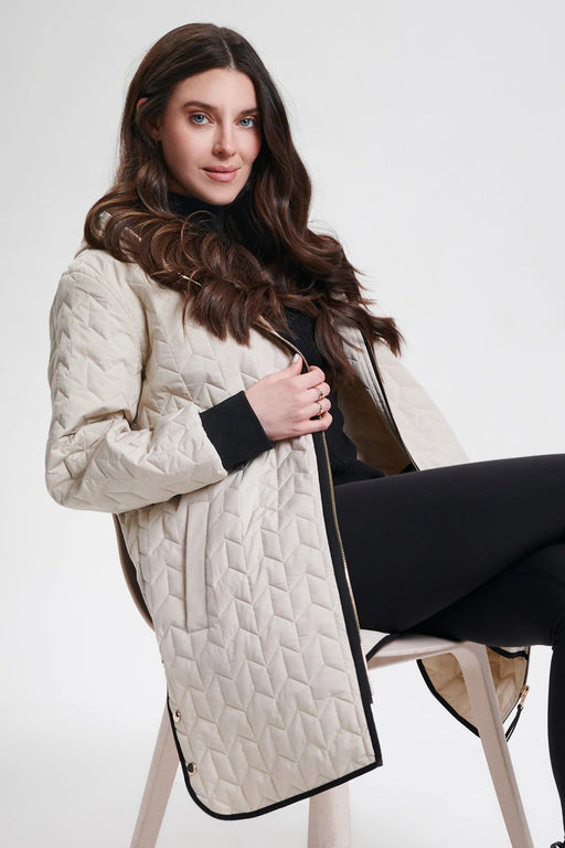 A woman seated on a chair wearing a long quilted cream-colored coat with hood from Nikki Jones