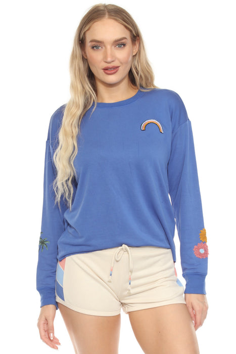 Woman wearing a blue pullover lounge top with rainbow, daisy, and palm tree embroidery
